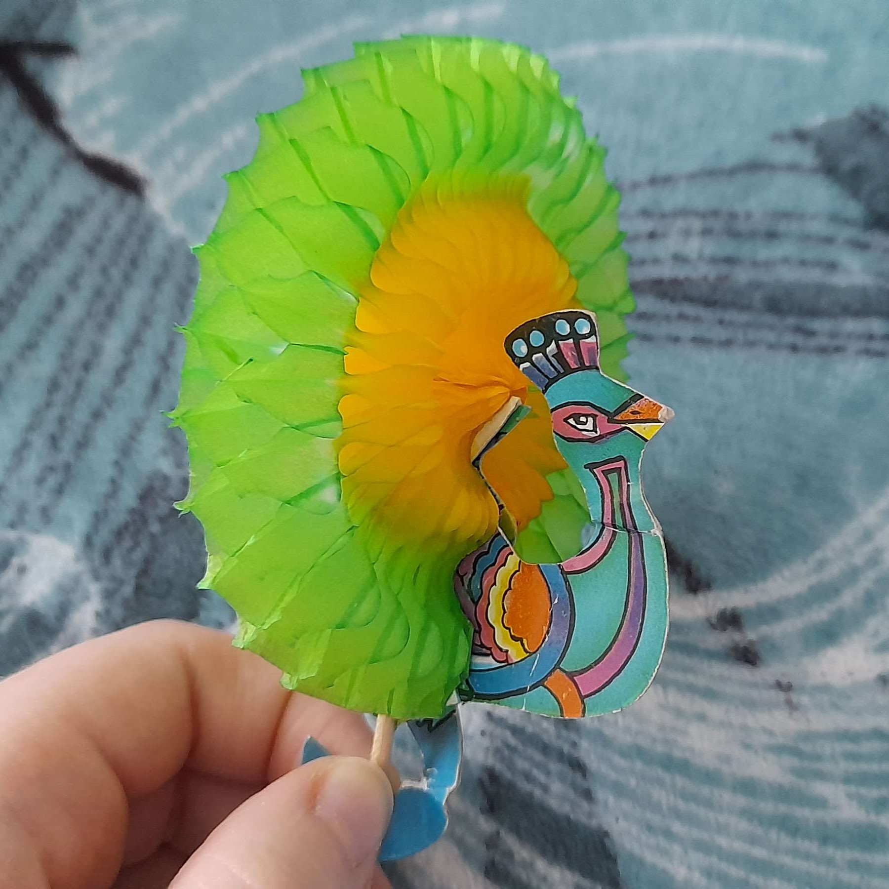 a paper peacock cocktail decoration thingy. the quote-unquote 'feathers' are spread out, revealing a green and yellow circle of paper as the tail