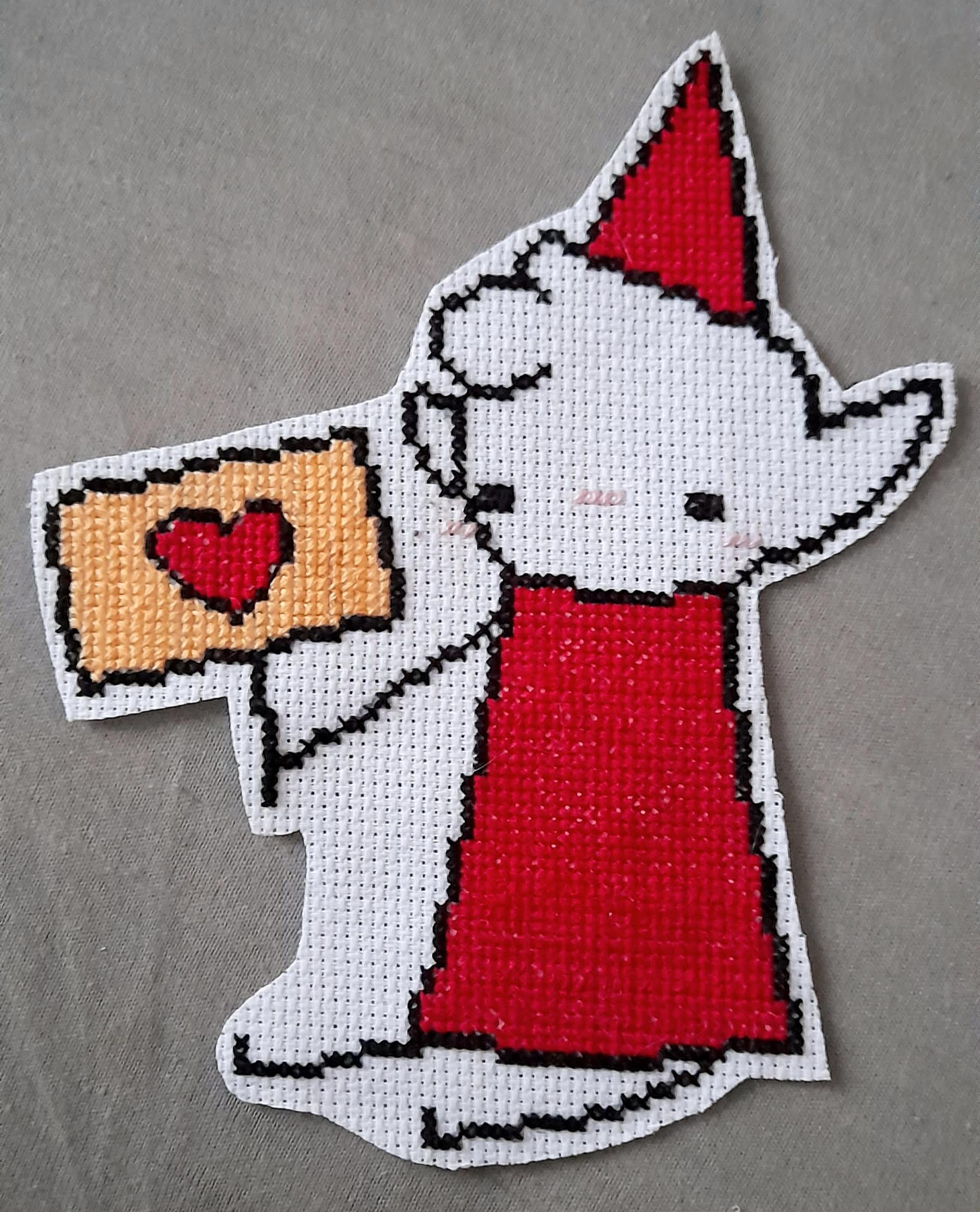 cross-stitch of alfur, a white elf wearing a red tunic and pointy hat. he is sitting down and holding up a sign with a heart on it