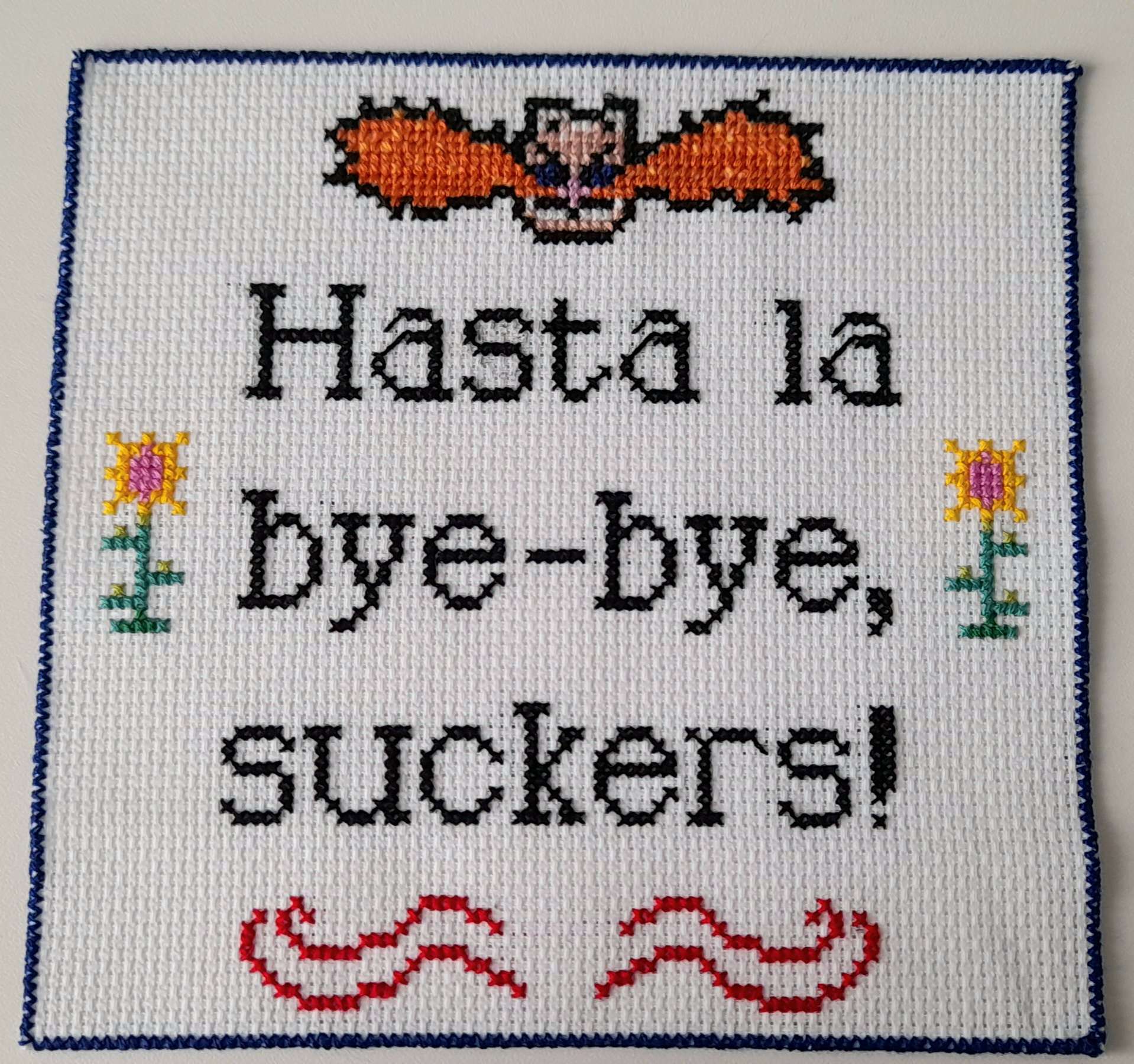 a cross-stitch piece showing text saying 'hasta la bye-bye, suckers!' with a small depiction of dr. eggman's head above it, red swirls below it, and yellow flowers on each side. the whole piece has a dark blue border