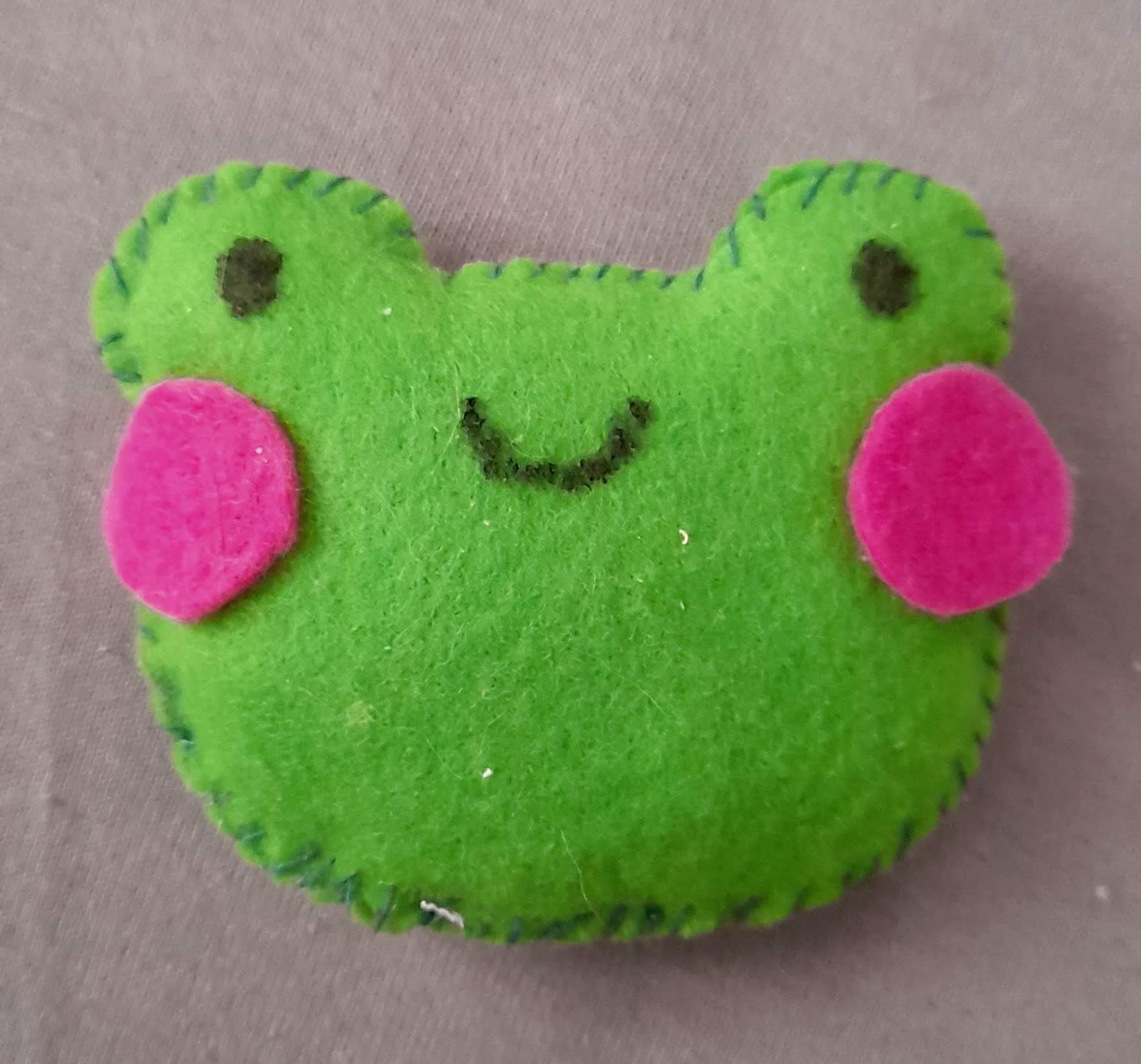 small plushie of a green smiling frog head with rosy cheeks